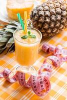 Diagonally left down a glass of pineapple smoothie with straws and mint leaf and open centimeter in the background pineapple and slice, on yellow checkered tablecloth. Close. Vertical. photo