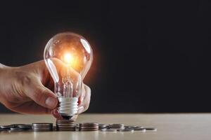 Hand holding light bulb and money, coins stack on wood table and on black background. Idea saving energy and accounting finance. ways to adjust your budget to this New Normal and save more concept photo
