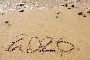 New Year 2026, inscription on the beach with sea wave. Number 2026 on the seashore, handwritten message on the sand. New Year concept. photo
