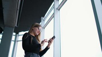 Beautiful woman doing photo of herself indoors. Attractive girl in glasses stands near the large windows and using mobile phone. View from below. video