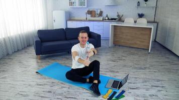 Young man exercising at home. Man is sitting on a mat on floor and holding balance with a bottle. Fitness at home during quarantine. video