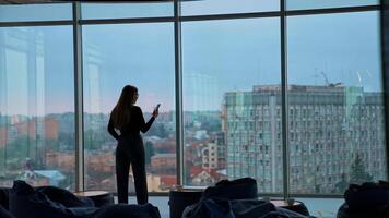 Young business woman with a phone. Rear view of a female in modern office near the large windows. Panoramic windows view with a city background. Back view. video
