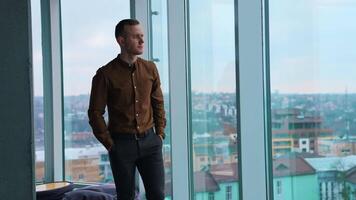 Full-length portrait of a businessman indoors. Tired young man walking along the office and stands by the large window with city view background. video