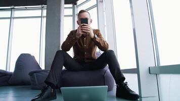 Young man with a phone in office. Handsome businessman sitting on a comfortable armchair and using a mobile phone on window background. Front view. video