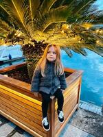 Little girl sits on a wooden tub with a palm tree with luminous garlands on the pier photo