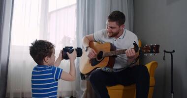 A father plays guitar while his son captures the moment, bonding video