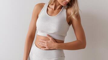 Woman in light sportswear gently touches her belly, symbolizing abdominal concern or diastasis recti awareness, standing in a neutral studio background. photo