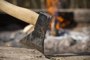 An axe is deeply embedded in a wood chopping block with a warm campfire blazing behind it. photo