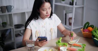 Handheld high angle view shot, Beautiful Asian woman delicious salad rolls while playing with mobile phone happily at home. Relax at home, Enjoy healthy food video