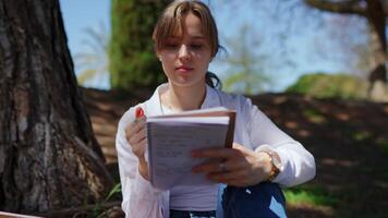 Female student sitting under leafy tree, writing notes while enjoying peaceful park atmosphere during bright sunny day, capturing learning moment with focused concentration video