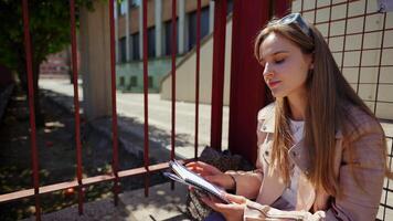 Focused college student reviewing study notes while sitting on low wall, metal fence and building backdrop creating urban academic scene during sunny day video