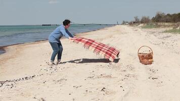 Woman spreads picnic plaid blanket at beach sand to have picnic on sea beach. Waves wash shore, sea coast nature, seascape. Outdoor recreation, picnic at beach. video