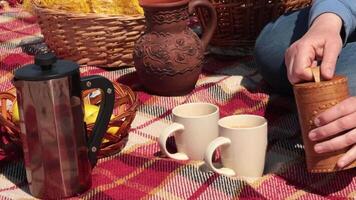 Person adds sugar from jar named Sugar into ceramic mugs and stir with spoon. Process of making coffee at picnic. Rustic style picnic still life with woven baskets on plaid blanket video