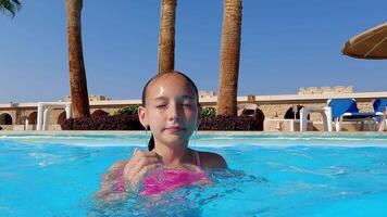 Young girl emerging from the sparkling turquoise water of a sunny resort swimming pool, eyes closed in blissful relaxation, surrounded by lush palm trees and vibrant tropical scenery video