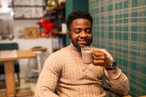 Young African American man enjoying hot chocolate at cafe. He is sitting at table indoor with eyes closed in satisfaction, smiling and relaxed after work. photo