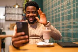 Young black man smiling and waving during a call on his smartphone, enjoying a conversation in warm cafe environment. photo