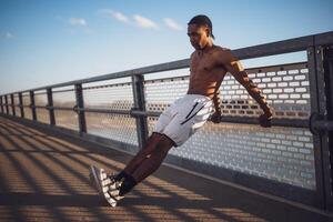 Shirtless, muscular African man working out on bridge in the city. He is practicing reverse push ups to build strength and endurance. photo