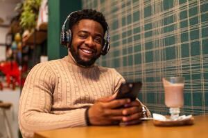 Cheerful African American man with headphones enjoying a break while sitting in a cafe, holding a smartphone and smiling at the camera. photo