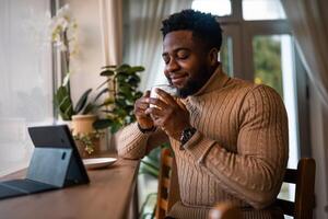 Young African man enjoying sipping tea and relaxing while working remotely with digital tablet from stylish coffee shop. photo