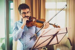 Young male violinist practicing at home with intense focus, wearing eyeglasses and holding violin in quiet room. photo
