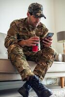 Depressed soldier wearing camouflage uniform sitting on sofa using smartphone and drinking energy drink photo