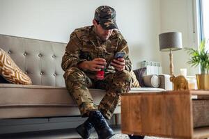 Depressed soldier wearing camouflage uniform sitting on sofa using smartphone and drinking soda photo