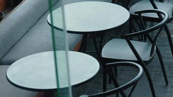 A close-up view of two round glass tables with black chairs in a modern cafe setting. The tables are empty, and there is a gray cushioned bench in the background video