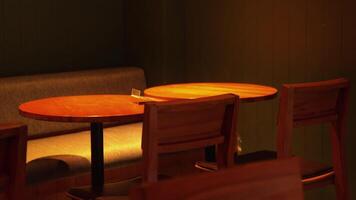 A cozy interior scene featuring two wooden tables and several wooden chairs. The lighting is warm, creating a relaxed atmosphere. A gray upholstered bench is visible in the background. video
