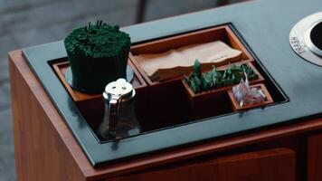 A wooden table with a black surface featuring various condiments and utensils. There are green chopsticks, napkins, and small containers with herbs and sugar. A glass jar is also present. video