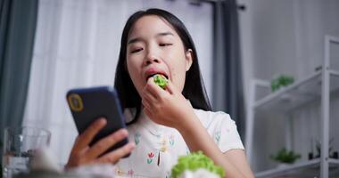 Handheld low angle view shot, Beautiful Asian woman delicious salad rolls while playing with mobile phone happily at home. Relax at home, Enjoy healthy food video