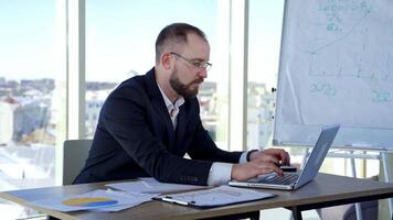 Serious male worker using laptop computer. Focused businessman working on laptop in office with large windows. Businessman in glasses typing on a computer. video
