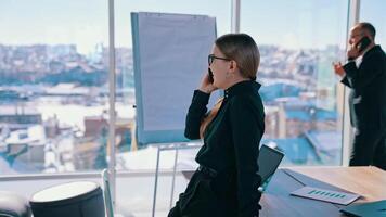 Business people talking on their phones indoors. Smiling woman having a conversation through the phone in modern office. Businessman near the window with city background. video
