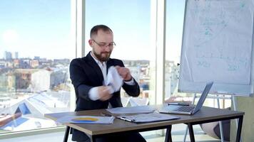 Nervous businessman in office. Irritated man entrepreneur sitting at table and tearing papers throwing them to the sides on the background of window. video