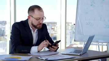Bearded businessman in glasses working in office. Serious office worker in suit sitting at the desk using the phone and talking. video