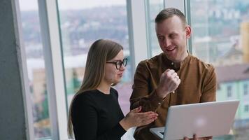 Happy business partners talk in office. Man and woman speaking about new solution of a project while standing together with a laptop on window city view background. video