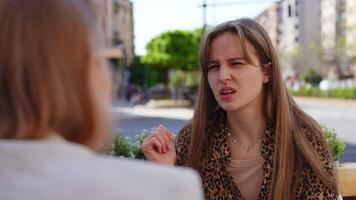 Young woman with long hair and a leopard print jacket is sitting at an outdoor bar table, gesturing and explaining something to a friend who is listening attentively video
