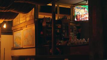 A cozy cafe interior featuring wooden shelves filled with various items, a television displaying a colorful scene, and maps on the wall. The warm lighting creates an inviting atmosphere. video