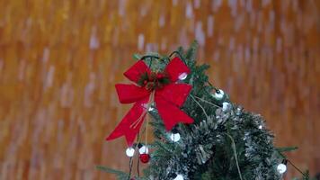A close-up of a Christmas tree top adorned with a red bow, pinecones, and white lights, set against a warm wooden background. video