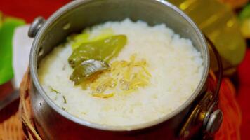 A close-up of a pot filled with fluffy white rice, garnished with green chilies and herbs. The pot is made of metal and has a rustic appearance, sitting on a woven mat. video