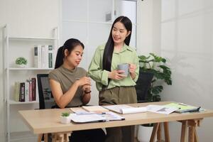 Portrait of Two colleagues are working together in an office. A young woman is standing holding a cup of coffee and exchanging ideas with another colleague who is sitting. photo