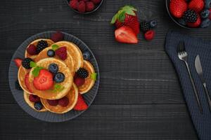 A flatlay of golden pancakes on a black wooden background, topped with fresh berries create a cozy and indulgent scene. photo