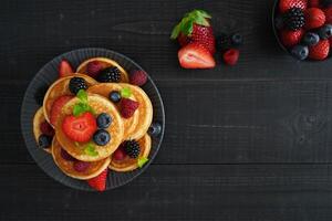 A flatlay of golden pancakes on a black wooden background, topped with fresh berries create a cozy and indulgent scene photo