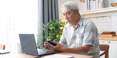 Senior man checking on mobile phone while working with laptop at home photo
