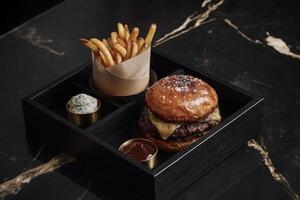 Black stained oak tray with tasty burger, fries and dipping cups of sauce on a dark background. photo
