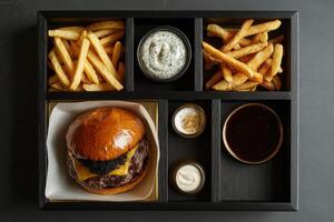 Black stained oak tray with tasty burger wrapped in paper, fries and dipping cups of sauce on a dark background. photo