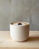 A matte white ceramic cup with iced coffee on a light birchwood surface. A single perfect coffee bean on the foam. photo