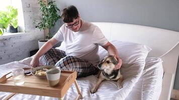 man in home clothes sitting on the bed using laptop working or playing games and playing with dog video