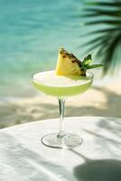 A glass filled with a frothy summer pineapple mint cocktail. The setting is a white onyx bar table against a beach photo