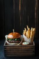 A wooden tray with tasty burger wrapped in unbleached parchment paper, the fries and a ceramic dipping cup of smoked sauce. photo