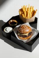 Black oak tray with tasty burger wrapped in paper, fries and dipping cups of sauce on a white background. photo